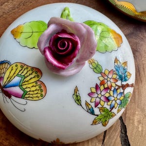 May include: A white porcelain lid with a pink rose, green leaves, a yellow and blue butterfly, and colorful flowers. The lid is decorated with gold accents.