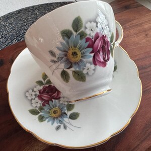May include: A white teacup and saucer with a floral design. The design features pink roses, blue flowers, and green leaves. The cup and saucer have a gold trim.