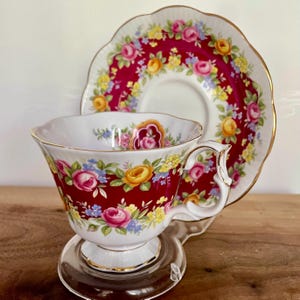 May include: A vintage teacup and saucer set. The teacup and saucer are decorated with a vibrant floral pattern of pink, yellow, and blue roses against a deep red background. The cup has a white interior and a gold trim.