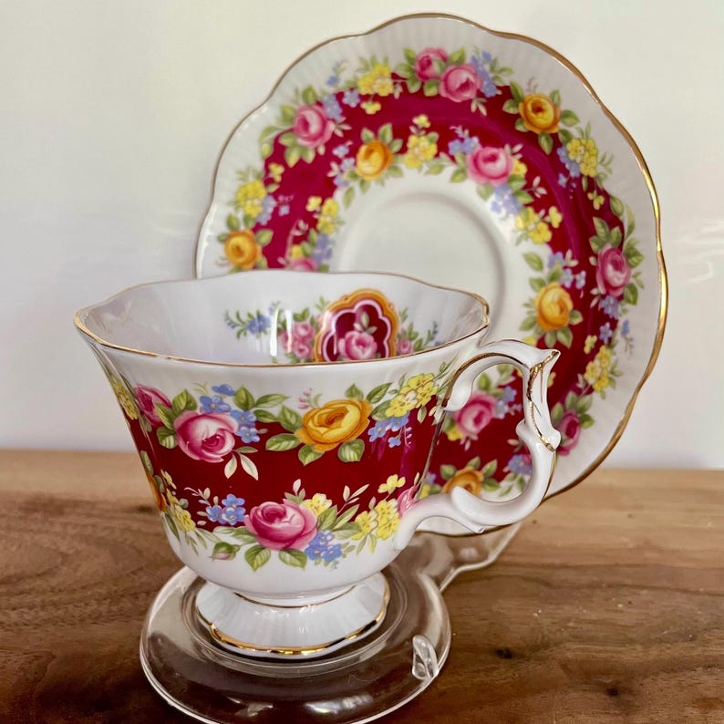 May include: A vintage teacup and saucer set. The teacup and saucer are decorated with a vibrant floral pattern of pink, yellow, and blue roses against a deep red background. The cup has a white interior and a gold trim.