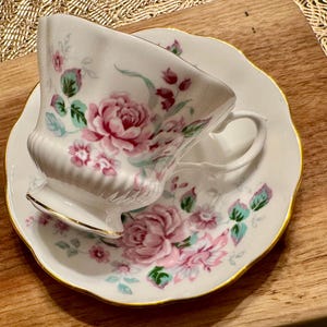 May include: A white teacup and saucer set with a floral pattern featuring pink and green roses. The set has a gold trim around the edges.