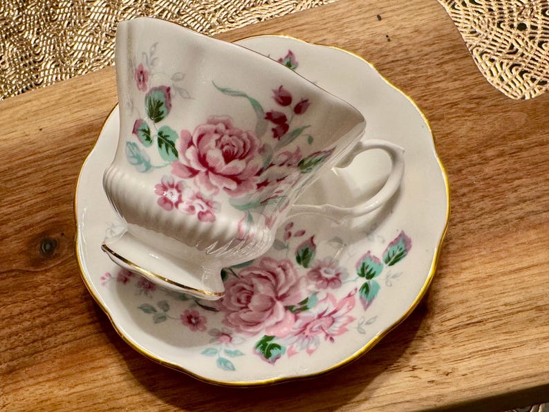 May include: A white teacup and saucer set with a floral pattern featuring pink and green roses. The set has a gold trim around the edges.