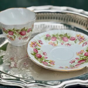 May include: A white teacup and saucer set with a floral pattern of pink roses and green leaves, with gold trim. The set is on a silver tray with an embossed floral design. A classic tea set.