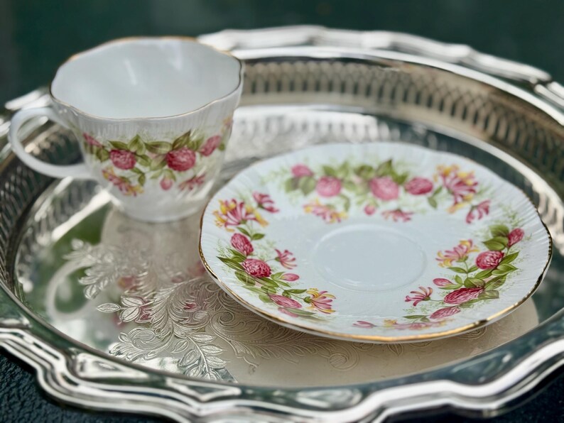 May include: A white teacup and saucer set with a floral pattern of pink roses and green leaves, with gold trim. The set is on a silver tray with an embossed floral design. A classic tea set.