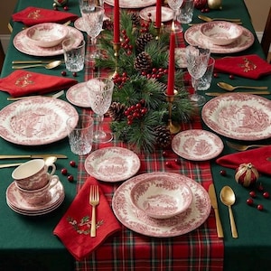 May include: A Christmas table setting with red and white china, gold cutlery, and red napkins. The centerpiece features pine boughs, berries, and red candles. The china has a scenic design, ideal for a festive meal.