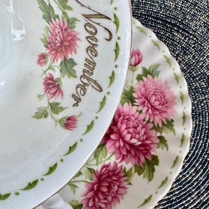 May include: A white teacup and saucer set with a floral design featuring pink chrysanthemums and green leaves. The word "November" is written on the cup. The set has a gold trim.