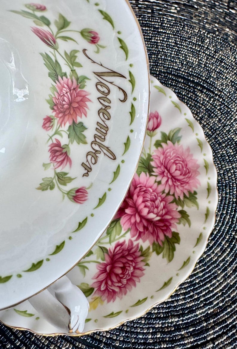 May include: A white teacup and saucer set with a floral design featuring pink chrysanthemums and green leaves. The word "November" is written on the cup. The set has a gold trim.