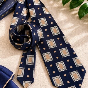 May include: Navy blue tie with a repeating pattern of tan and white squares on a dark background. The tie is knotted and lies next to a matching pocket square and a pen on a book titled "Timeless Style."