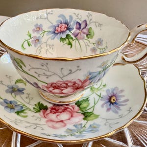 Pu&ograve; includere: Un set vintage di tazza e piattino da t&egrave;. La tazza e il piattino in porcellana bianca sono adornati da un motivo floreale nei toni del rosa, blu e viola, con bordi dorati. La tazza ha un manico curvo.
