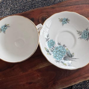 May include: A white teacup and saucer set with a blue floral design. The saucer has a scalloped edge and the teacup has a gold rim.