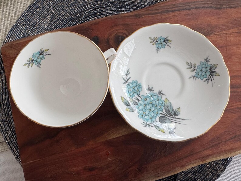 May include: A white teacup and saucer set with a blue floral design. The saucer has a scalloped edge and the teacup has a gold rim.
