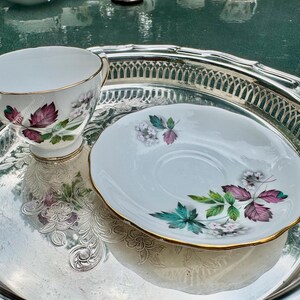 May include: A white teacup and saucer set with a floral design in shades of purple, green, and white, with gold trim. The set sits on a silver serving tray with a decorative edge. A classic tea set.
