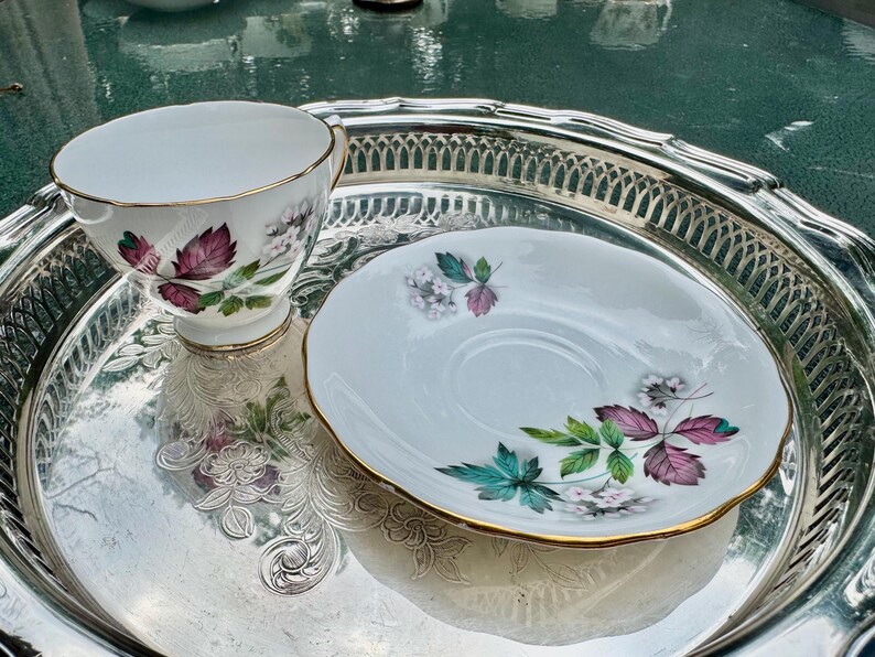 May include: A white teacup and saucer set with a floral design in shades of purple, green, and white, with gold trim. The set sits on a silver serving tray with a decorative edge. A classic tea set.
