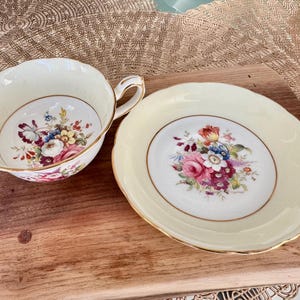 May include: A vintage teacup and saucer set with a floral pattern. The cup and saucer are both cream colored with a gold trim. The floral pattern features pink, red, yellow, and blue flowers.