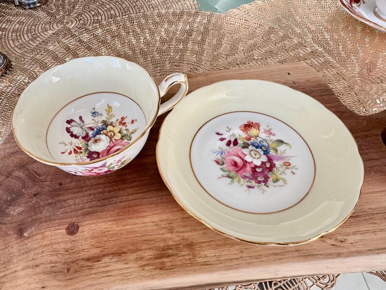 May include: A vintage teacup and saucer set with a floral pattern. The cup and saucer are both cream colored with a gold trim. The floral pattern features pink, red, yellow, and blue flowers.