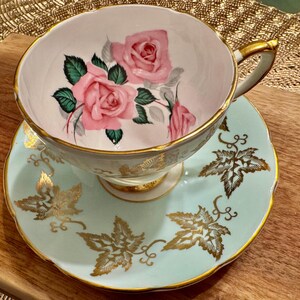 May include: A vintage teacup and saucer set with a delicate floral design. The cup features pink roses and green leaves on a white background, while the saucer is a light blue with gold leaf accents.