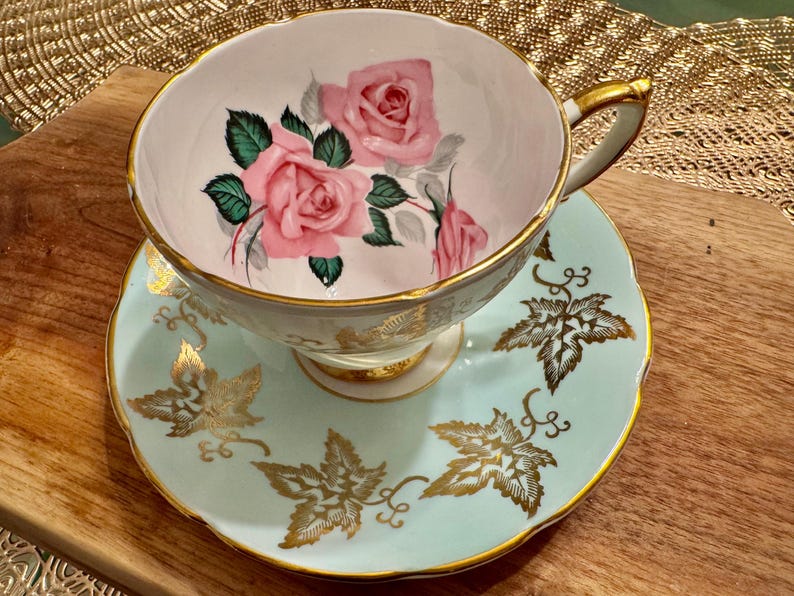 May include: A vintage teacup and saucer set with a delicate floral design. The cup features pink roses and green leaves on a white background, while the saucer is a light blue with gold leaf accents.