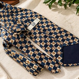 May include: A necktie featuring a repeating geometric pattern of dark blue squares alternating with tan and brown floral-like motifs. The tie includes a white label with the text GAP printed in black on the back side and a solid dark blue lining.