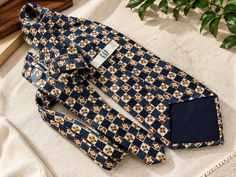 May include: A necktie featuring a repeating geometric pattern of dark blue squares alternating with tan and brown floral-like motifs. The tie includes a white label with the text GAP printed in black on the back side and a solid dark blue lining.