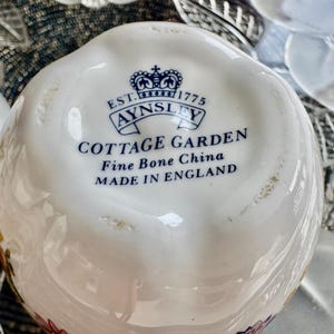 K&ouml;nnte beinhalten: Nahaufnahme der Unterseite einer wei&szlig;en Teetasse aus Keramik mit einem blauen und wei&szlig;en Blumenmuster. Auf der Unterseite der Tasse steht der Schriftzug "EST. 1775 AYNSLEY COTTAGE GARDEN Fine Bone China MADE IN ENGLAND".