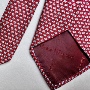 Puede incluir: Una corbata roja con un patr&oacute;n repetitivo de peque&ntilde;os elefantes blancos y azul claro. La parte posterior de la corbata muestra un forro rojo oscuro con el texto bordado Salvatore Ferragamo repetido en la tela.