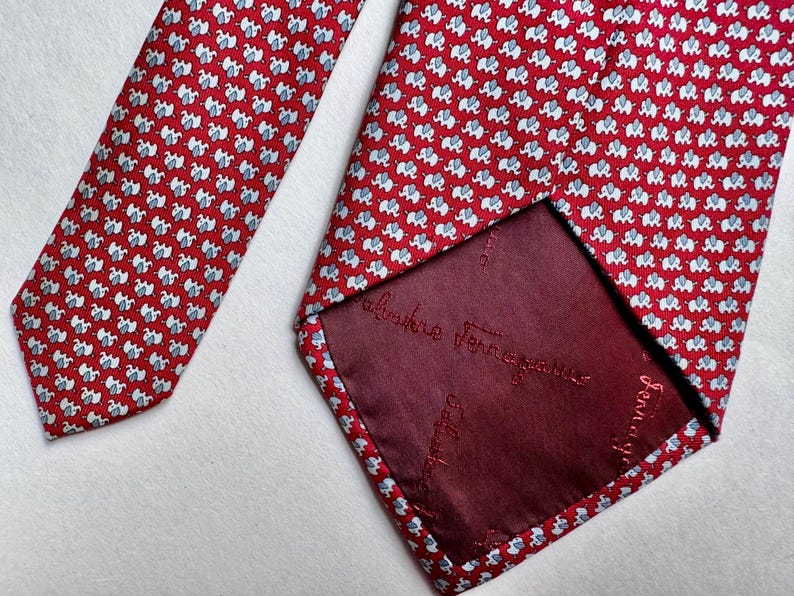 Puede incluir: Una corbata roja con un patr&oacute;n repetitivo de peque&ntilde;os elefantes blancos y azul claro. La parte posterior de la corbata muestra un forro rojo oscuro con el texto bordado Salvatore Ferragamo repetido en la tela.