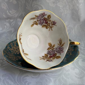 Może przedstawiać: Biały porcelanowy zestaw filiżanki ze spodkiem o falistym brzegu ze złotym wykończeniem. Wnętrze filiżanki zdobią motywy fioletowych r&oacute;ż z zielonymi liśćmi. Spodek jest w kolorze morskim z misternym złotym wzorem filigranowym na obrzeżach.