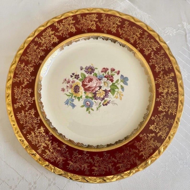 May include: A vintage decorative plate with a cream-colored center featuring a floral bouquet in shades of pink, purple, yellow, and blue. The rim is a deep burgundy with gold floral detailing and a gold edge.