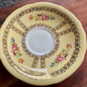 May include: A round ceramic saucer with a pale yellow background, featuring a central white circle. The saucer is decorated with a gold leaf garland pattern and small clusters of pink, yellow, and blue flowers arranged around the center.
