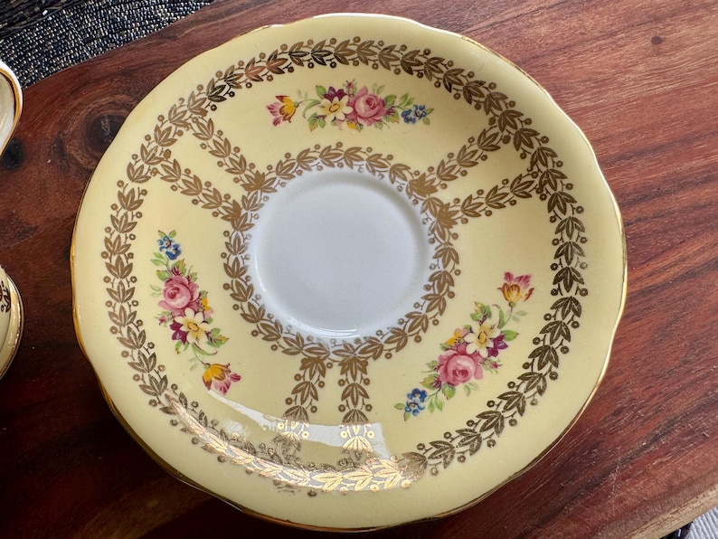 May include: A round ceramic saucer with a pale yellow background, featuring a central white circle. The saucer is decorated with a gold leaf garland pattern and small clusters of pink, yellow, and blue flowers arranged around the center.