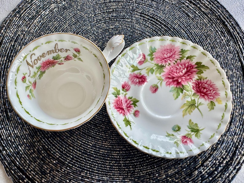 May include: A white teacup and saucer set with a floral design featuring pink flowers and green leaves. The teacup has the word "November" written on it. The set has gold trim.