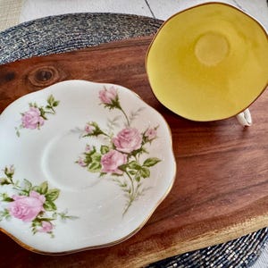 Może przedstawiać: Zestaw filiżanki i spodeczka do herbaty z białej porcelany z kwiatowym wzorem przedstawiającym r&oacute;żowe r&oacute;że i zielone liście. Spodeczek ma falisty brzeg, a filiżanka do herbaty ma ż&oacute;łte wnętrze.