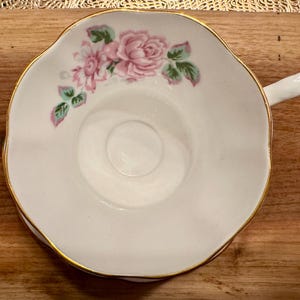 May include: A white teacup with a gold rim and a floral design featuring pink roses and green leaves.