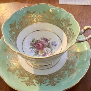 Peut inclure: Un ensemble tasse et soucoupe en porcelaine avec une bordure vert menthe p&acirc;le orn&eacute;e de filigranes dor&eacute;s complexes. Le centre de la tasse pr&eacute;sente un bouquet floral de roses roses, de violettes violettes et de marguerites blanches.