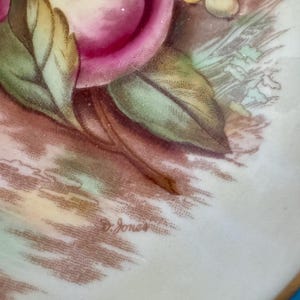May include: Close-up of a decorative plate with a floral design. The plate features pink roses, green leaves, and other floral elements. The artist's signature, "D. Jones", is visible on the plate's surface. The plate has a gold rim.