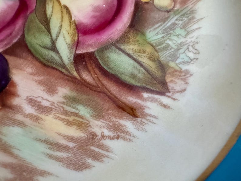 May include: Close-up of a decorative plate with a floral design. The plate features pink roses, green leaves, and other floral elements. The artist's signature, "D. Jones", is visible on the plate's surface. The plate has a gold rim.