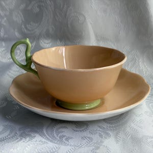 May include: A peach-colored teacup and saucer set featuring a scalloped rim and a light green handle. The teacup has a matching light green base, while the saucer has a white underside. The ceramic surface has a smooth, glossy finish throughout.