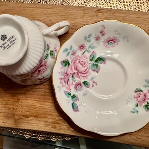 May include: A white teacup and saucer with a floral pattern. The teacup has a gold rim and the saucer has a scalloped edge. The teacup has the words "Royal Albert Bone China England" printed on the bottom.