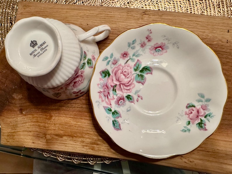 May include: A white teacup and saucer with a floral pattern. The teacup has a gold rim and the saucer has a scalloped edge. The teacup has the words "Royal Albert Bone China England" printed on the bottom.