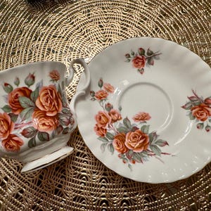 Pode incluir: Um conjunto de ch&aacute; branco com x&iacute;cara e pires com um design floral com rosas cor-de-rosa e laranja. O conjunto &eacute; decorado com uma delicada borda dourada.