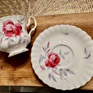 May include: A white teacup and saucer with a pink rose design. The cup has a scalloped edge and a handle. The saucer has a scalloped edge and a floral design.