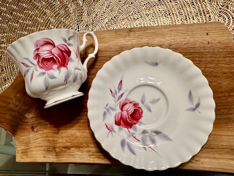 May include: A white teacup and saucer with a pink rose design. The cup has a scalloped edge and a handle. The saucer has a scalloped edge and a floral design.