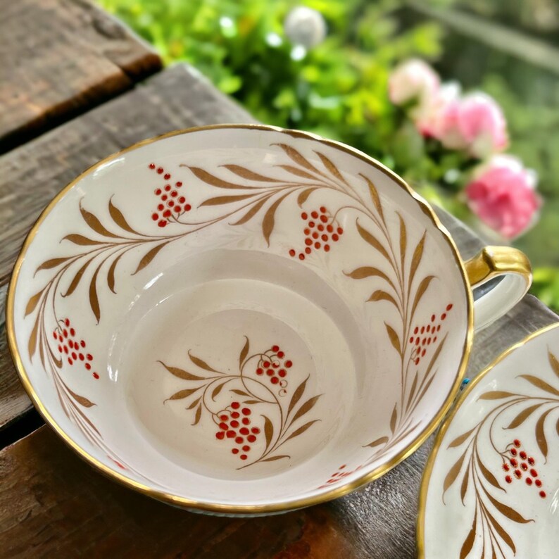 Pu&ograve; includere: Un set di tazza e piattino bianchi con finiture dorate e un motivo decorativo di foglie dorate e bacche rosse. La tazza ha un manico dorato. Il set &egrave; posto su una superficie di legno.