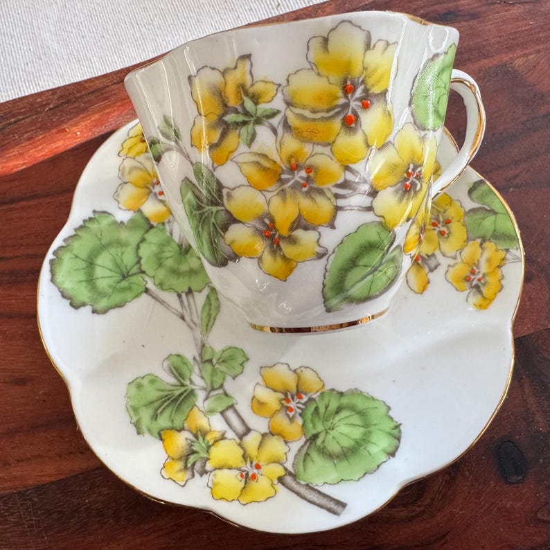 May include: A white teacup and saucer with a floral design. The cup and saucer feature yellow flowers with green leaves on a white background. The cup has a gold rim.