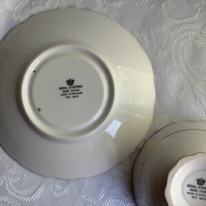 May include: Two off-white bone china pieces with scalloped edges. The underside of each piece features a black stamp with a crown icon and the text: ROYAL STAFFORD BONE CHINA MADE IN ENGLAND EST 1845. The pieces have a smooth, glossy finish.