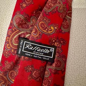 May include: A red necktie featuring a multi-colored paisley pattern in shades of gold, brown, and blue. A black label is stitched onto the fabric, displaying the text Raffaello Roma, Made in Italy, and 100% Seta-Silk in white lettering.