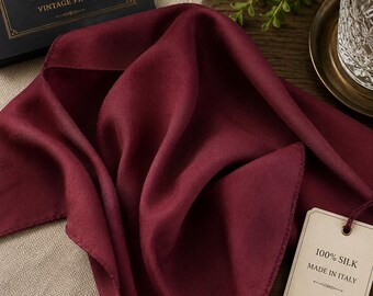 Puede incluir: Un pañuelo cuadrado de seda en color burdeos intenso con un acabado liso y mate. Una etiqueta de papel beige sujeta por un cordel indica 100% SILK MADE IN ITALY. El pañuelo está drapeado en pliegues suaves, mostrando su color sólido y rico.