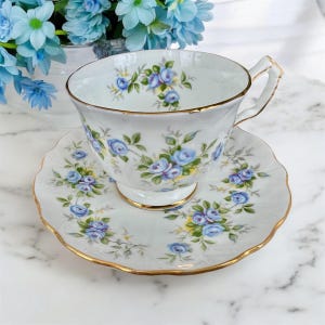 Pu&ograve; includere: Una tazza da t&egrave; e un piattino bianchi con un delicato motivo floreale blu e un bordo dorato. Il motivo floreale presenta rose blu e foglie verdi.