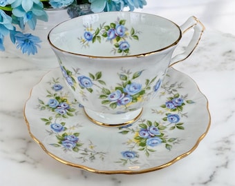 May include: A white teacup and saucer with a delicate blue floral pattern and gold trim. The floral design features blue roses and green leaves.