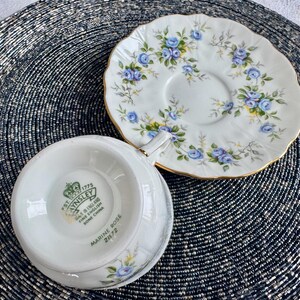 Pu&ograve; includere: Una tazzina da t&egrave; e un piattino bianchi con un motivo floreale blu e verde. Il piattino ha un bordo dorato. La tazzina da t&egrave; ha le parole "Aynsley Est. 1775 Fine Bone China Made in England" e "Marine Rose 2872" stampate sul fondo.